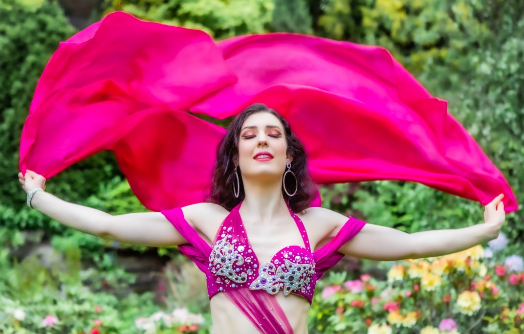 upper body photo of Rachael with arms outstretched, wearing a bright pink costume and holding a pink silk veil behind her