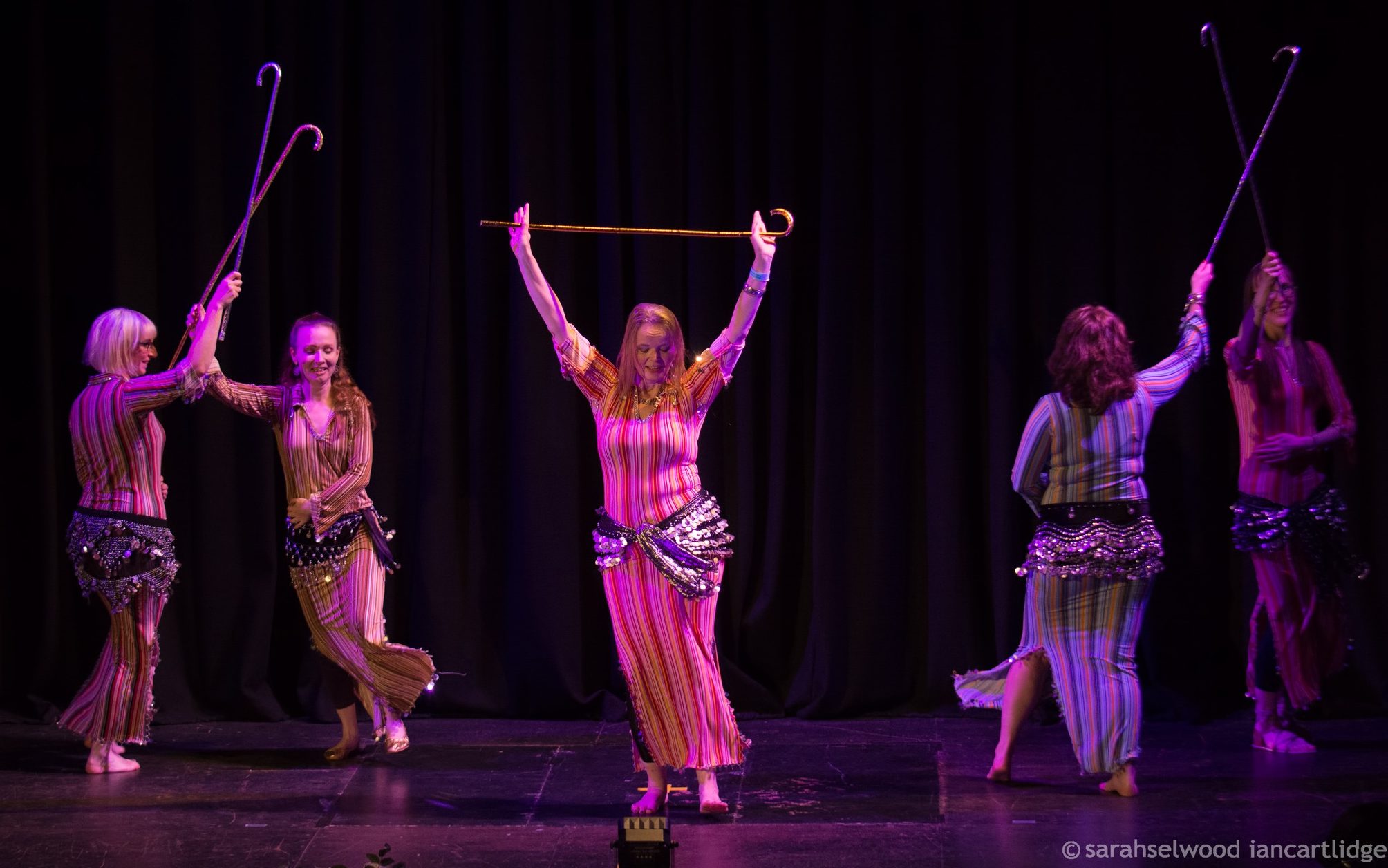 Five dancers on a stage, in front of dark curtains. They are wearing colourful striped dressed and coin scarves, holding sticks, and smiling