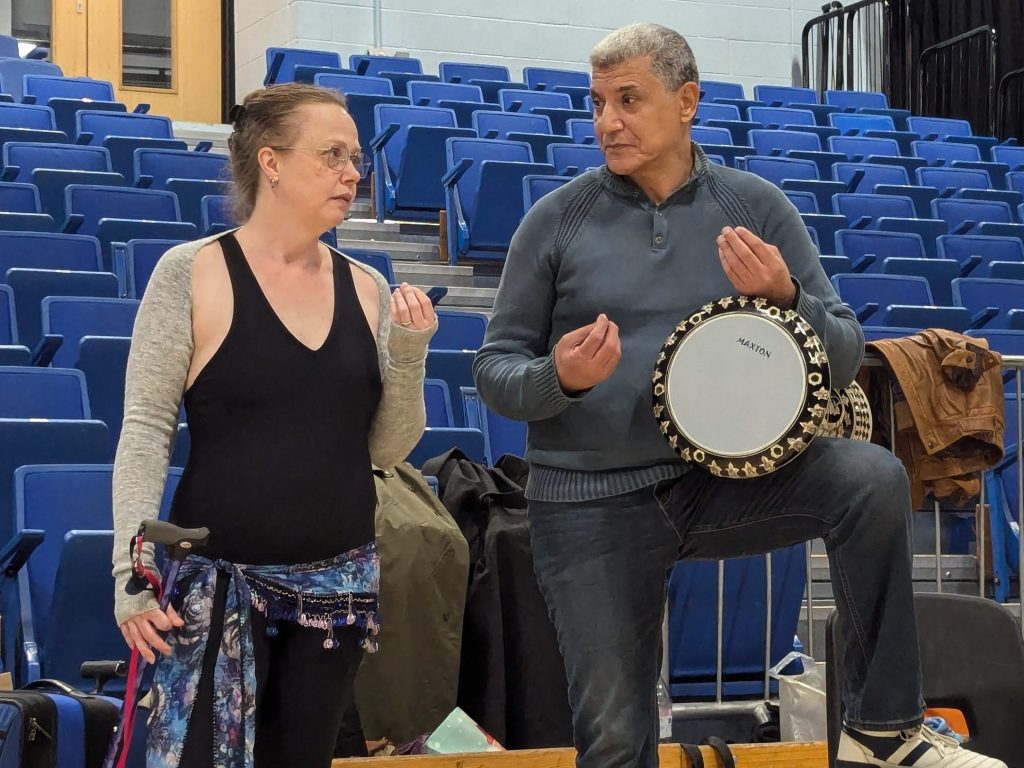 Dance researcher Nisaa stands on the left, and Egyptian musician Reda Henkesh stands on the right. Reda has a drum resting on his knee. Both are making a "slow down" hand gesture.