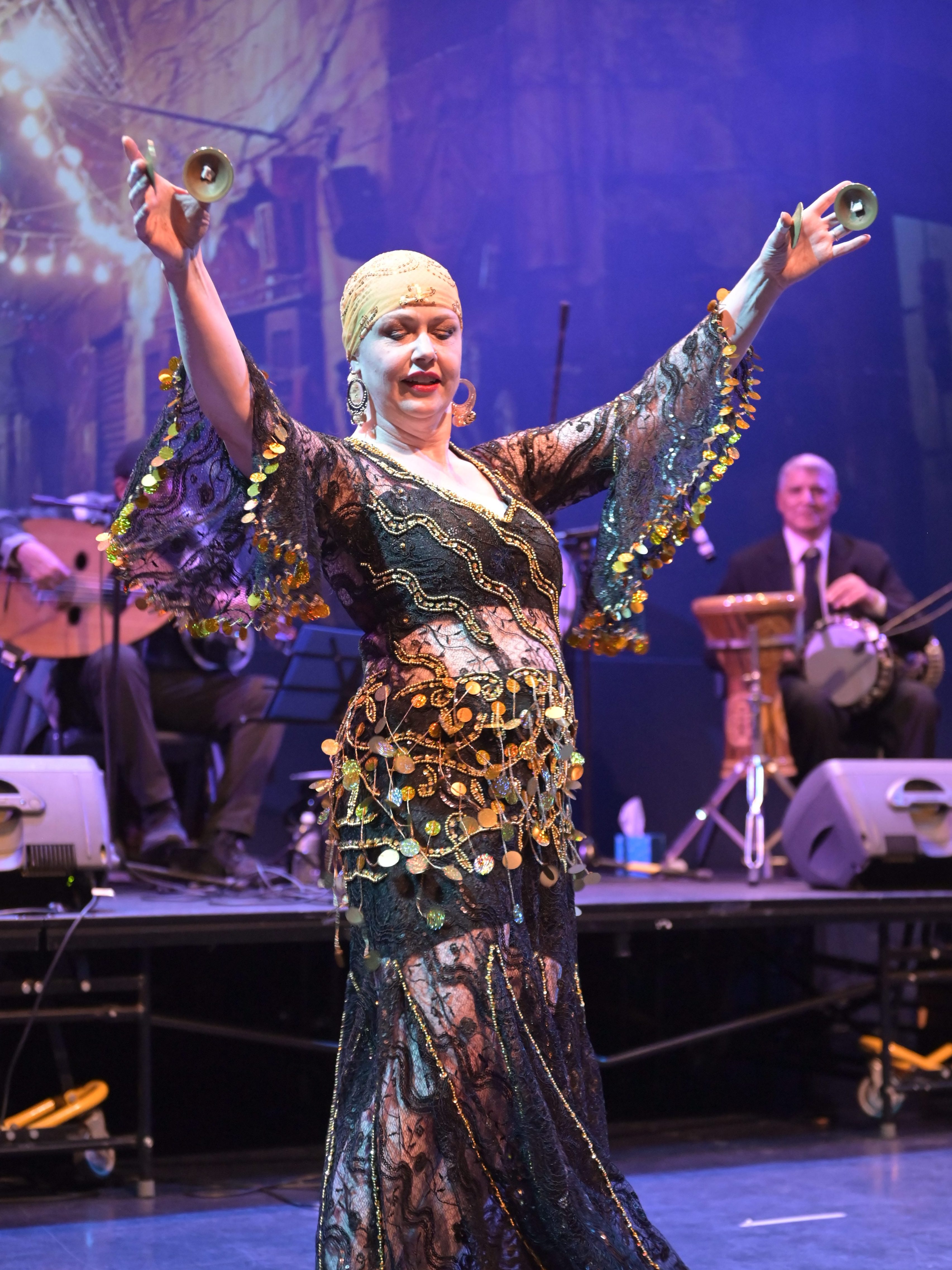 Nisaa is playing finger cymbals with her arms raised, wearing a black lace dress with gold sequins. 